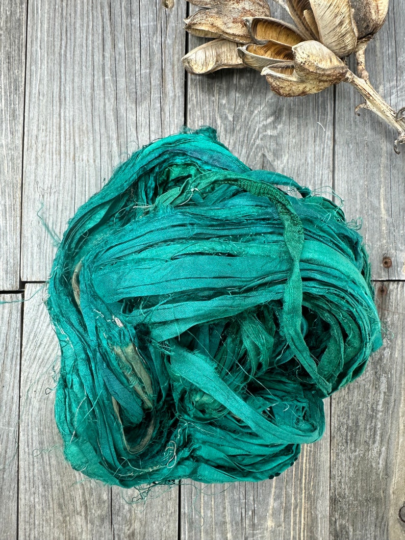 May include: A bundle of teal blue silk ribbon, also known as silk ribbon yarn, is shown on a wooden surface. The ribbon is made of thin strips of silk that have been twisted together.