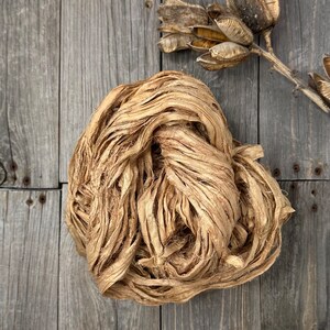 May include: A bundle of natural beige silk ribbon, also known as silk yarn or silk thread, is shown on a wooden surface. The silk ribbon is made from raw silk and is perfect for crafting and decorating.