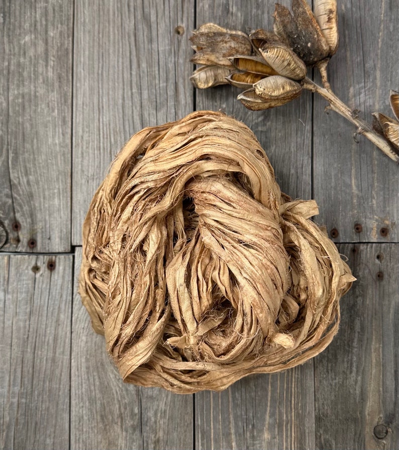 May include: A bundle of natural beige silk ribbon, also known as silk yarn or silk thread, is shown on a wooden surface. The silk ribbon is made from raw silk and is perfect for crafting and decorating.