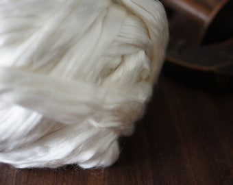 Pode incluir: Um close-up de uma grande bola branca de fibras de seda crua. As fibras são macias e fofas, e estão dispostas em uma massa solta e emaranhada.