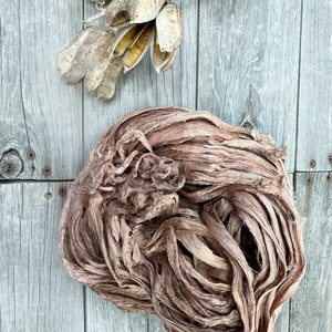 May include: A bundle of brown silk ribbon, with a slightly pink hue, is arranged in a loose, circular shape on a weathered wooden surface. The ribbon is made of thin, delicate strands that are twisted and knotted together.