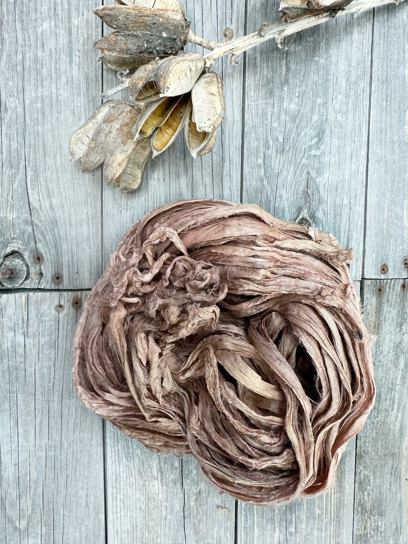 May include: A bundle of brown silk ribbon, with a slightly pink hue, is arranged in a loose, circular shape on a weathered wooden surface. The ribbon is made of thin, delicate strands that are twisted and knotted together.