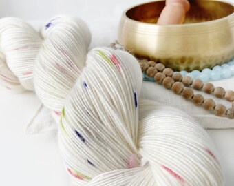 Puede incluir: Un ovillo de lana blanquecina con motas coloridas, junto a un cuenco tibetano dorado con un percutor de madera y hebras de cuentas. La lana tiene detalles rosas, azules y verdes. Los artículos están dispuestos sobre una superficie blanca.