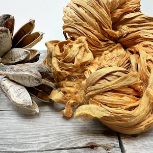 May include: A close-up of a bundle of golden silk ribbon, with a natural, rustic texture. The ribbon is arranged in a loose, organic shape, with some strands flowing freely. The ribbon is displayed on a light wood surface.