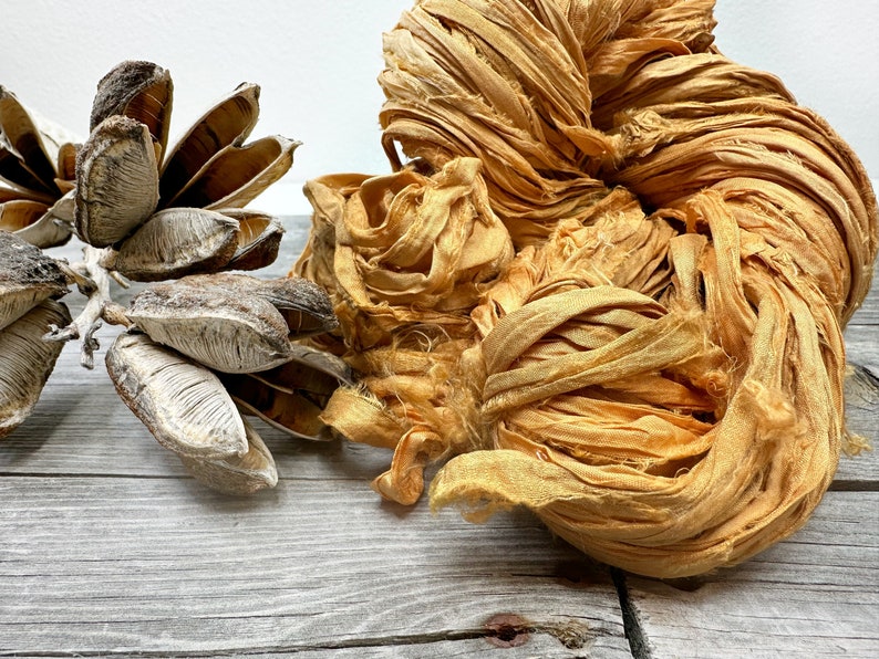 May include: A close-up of a bundle of golden silk ribbon, with a natural, rustic texture. The ribbon is arranged in a loose, organic shape, with some strands flowing freely. The ribbon is displayed on a light wood surface.