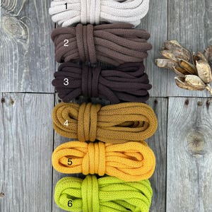 May include: Six bundles of thick, soft cotton rope in various colors: off-white, brown, dark brown, mustard yellow, bright yellow, and lime green. The ropes are neatly coiled and ready for crafting or decorative use. The background is a weathered wooden surface.