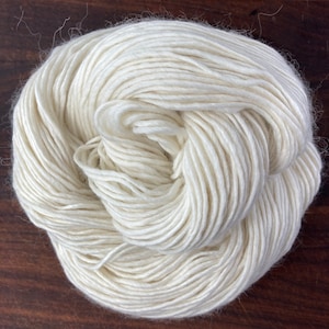 May include: A skein of white yarn, made from natural fibers, is shown on a wooden surface. The yarn is soft and fluffy, and it is perfect for knitting or crocheting.