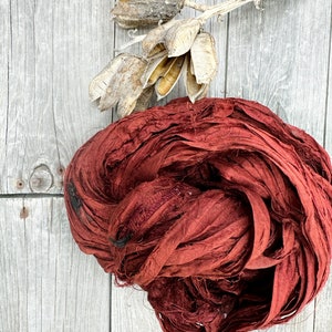 May include: A bundle of maroon silk ribbon, also known as silk ribbon yarn, on a wooden surface. The ribbon is made of thin strips of silk fabric that have been twisted together.
