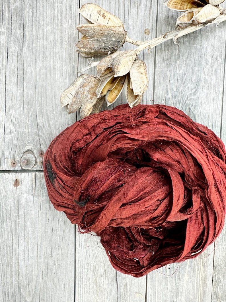 May include: A bundle of maroon silk ribbon, also known as silk ribbon yarn, on a wooden surface. The ribbon is made of thin strips of silk fabric that have been twisted together.