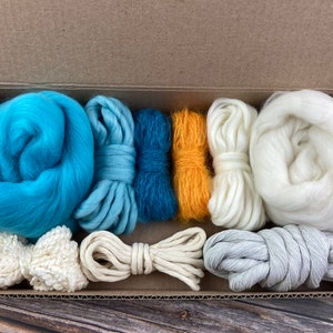 Puede incluir: Una caja llena de diferentes tonos de hilo azul, blanco y amarillo, junto con un lazo blanco de ganchillo y un trozo de cuerda natural. El hilo está dispuesto en una fila ordenada, listo para un proyecto de artesanía.