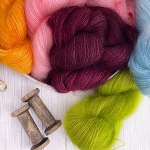 May include: Assortment of colourful yarn skeins in shades of orange, pink, burgundy, green, and blue. The yarn is arranged in a bowl and on a white wooden surface. Two wooden spools are also shown.