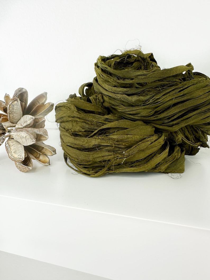 May include: A pile of olive green silk ribbon, also known as  silk ribbon yarn, on a white surface.
