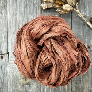 May include: A close-up of a bundle of brown silk ribbon. The ribbon is made of thin strips of silk that have been frayed and twisted together. The ribbon is lying on a wooden surface.