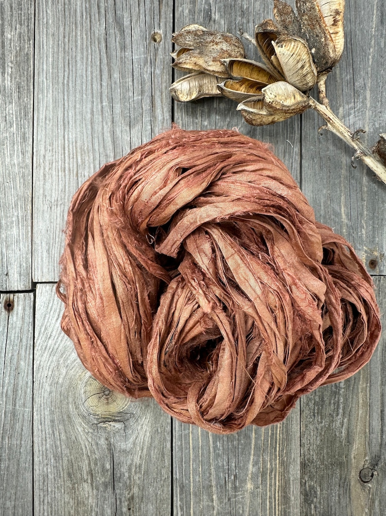 May include: A close-up of a bundle of brown silk ribbon. The ribbon is made of thin strips of silk that have been frayed and twisted together. The ribbon is lying on a wooden surface.