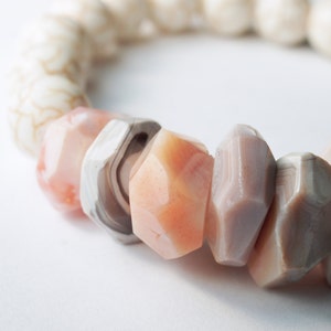 May include: A bracelet made of white, pink, and gray gemstone beads. The beads are faceted and have a natural, organic look.