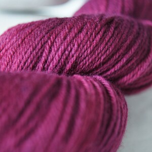 Puede incluir: Primer plano de un ovillo de hilo de color burdeos oscuro. El hilo es suave y esponjoso, con un brillo sutil.