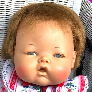 May include: A vintage baby doll with blue eyes, rosy cheeks, and a parted brown wig. The doll is wearing a white and red striped sleeper with a floral pattern and a white lace trim.