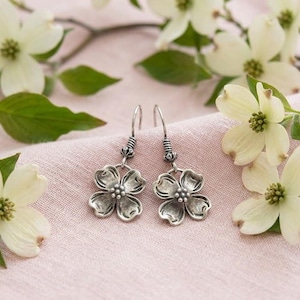 May include: A pair of silver-toned flower earrings with a hook closure. The earrings are displayed on a pink cloth, surrounded by white dogwood flowers and green leaves. The earrings have a detailed floral design.