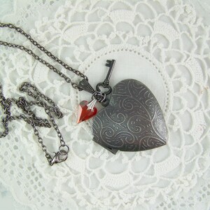 May include: A silver heart-shaped locket necklace with a key charm and two red crystal accents. The necklace is on a white lace background.