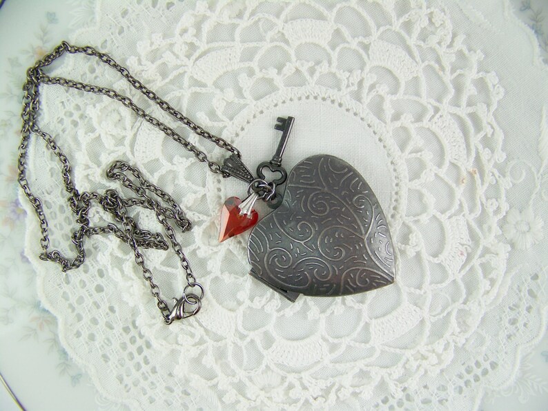 May include: A silver heart-shaped locket necklace with a key charm and two red crystal accents. The necklace is on a white lace background.