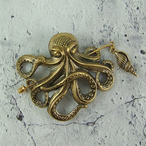 K&ouml;nnte beinhalten: Eine goldfarbene Brosche in Form eines Kraken mit einem Muschel-Anh&auml;nger. Der Krake hat detaillierte Tentakel und einen strukturierten K&ouml;rper. Die Brosche ist an einer Nadel befestigt.