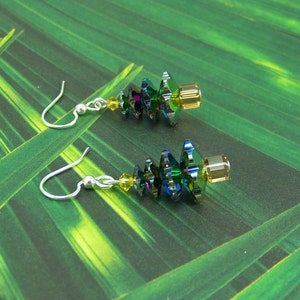 May include: A pair of dangle earrings featuring iridescent green and blue crystal beads with yellow accents. The earrings have silver-colored fish hook ear wires and are set against a green background.