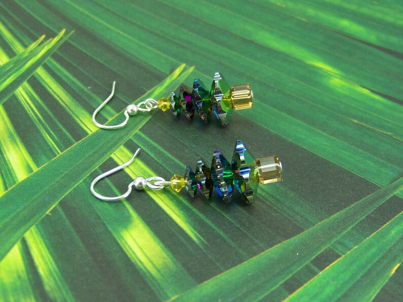 May include: A pair of dangle earrings featuring iridescent green and blue crystal beads with yellow accents. The earrings have silver-colored fish hook ear wires and are set against a green background.