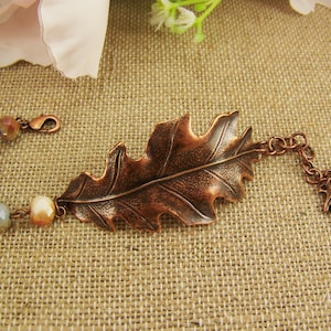 May include: A copper-toned bracelet featuring a large central oak leaf charm, a series of multi-colored faceted glass beads on one side, and a chain with a small acorn charm and a tiny leaf charm on the other, secured with a lobster claw clasp.