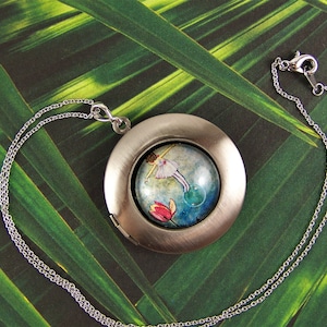 May include: A silver locket necklace with a round, glass-like pendant featuring a watercolor painting of a girl blowing bubbles. The pendant is surrounded by a silver frame.