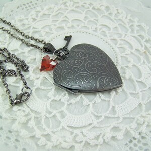 May include: A silver heart-shaped locket necklace with a key charm and two red crystal accents. The necklace is on a white lace background.