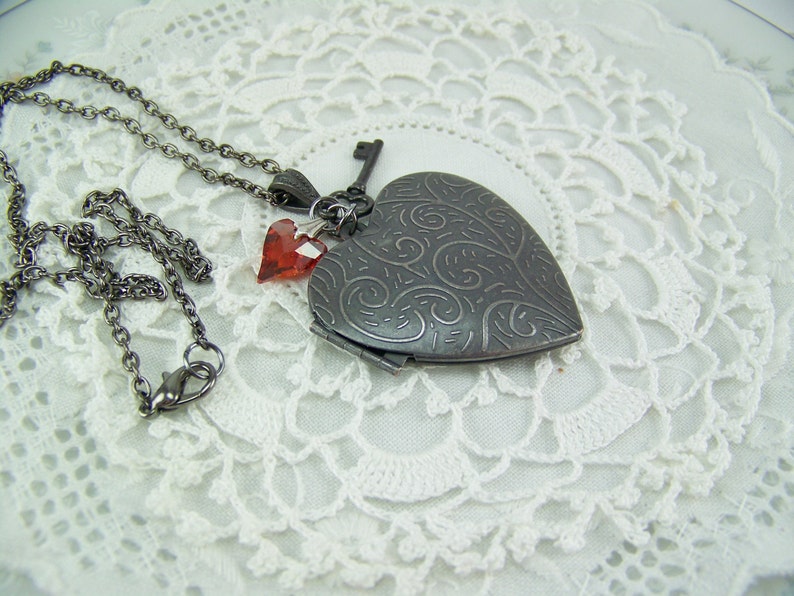May include: A silver heart-shaped locket necklace with a key charm and two red crystal accents. The necklace is on a white lace background.