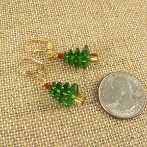 Peut inclure: Une paire de boucles d'oreilles en or avec des breloques en verre vert en forme de sapin de No&euml;l. Les boucles d'oreilles ont une perle rouge sur le fil au-dessus du charme.