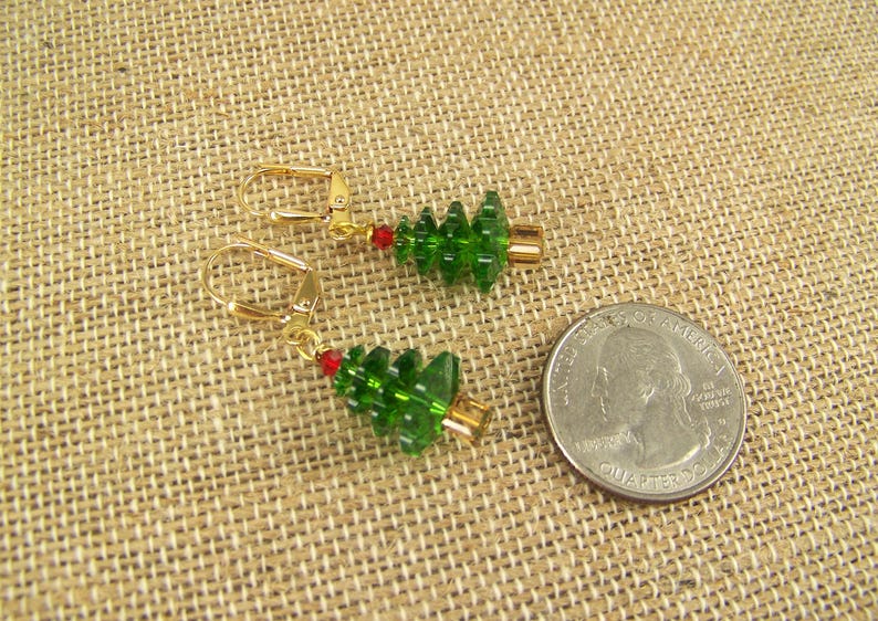 Peut inclure: Une paire de boucles d'oreilles en or avec des breloques en verre vert en forme de sapin de No&euml;l. Les boucles d'oreilles ont une perle rouge sur le fil au-dessus du charme.