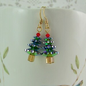 May include: A pair of Christmas tree earrings. Each earring features a gold-colored hook, a red bead at the top, and a tree made of iridescent green and blue faceted beads. The base of each tree is a gold-colored cube.