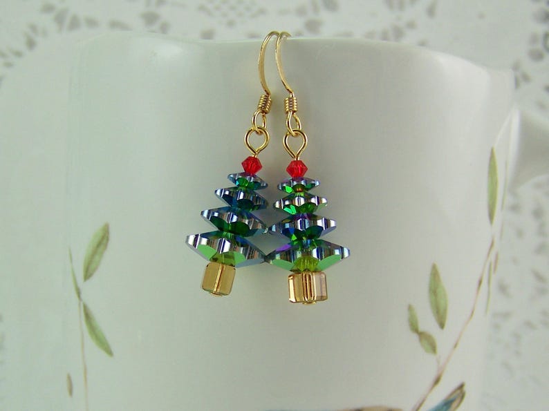May include: A pair of Christmas tree earrings. Each earring features a gold-colored hook, a red bead at the top, and a tree made of iridescent green and blue faceted beads. The base of each tree is a gold-colored cube.