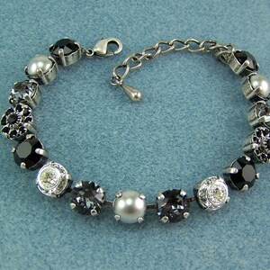 Puede incluir: Una pulsera que presenta una serie de piedras redondas facetadas en negro, gris y transparente intercaladas con perlas blancas lisas, todo montado en engastes de metal plateado. Incluye un cierre de mosquet&oacute;n y una cadena de extensi&oacute;n ajustable.