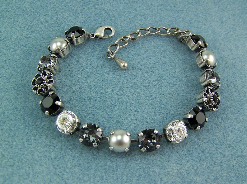 Puede incluir: Una pulsera que presenta una serie de piedras redondas facetadas en negro, gris y transparente intercaladas con perlas blancas lisas, todo montado en engastes de metal plateado. Incluye un cierre de mosquet&oacute;n y una cadena de extensi&oacute;n ajustable.