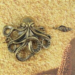 K&ouml;nnte beinhalten: Eine goldfarbene Brosche in Form eines Kraken mit einem detaillierten Design und einem Anh&auml;nger in Form einer Muschel. Die Brosche ist ein dekoratives Accessoire f&uuml;r Kleidung oder Taschen.