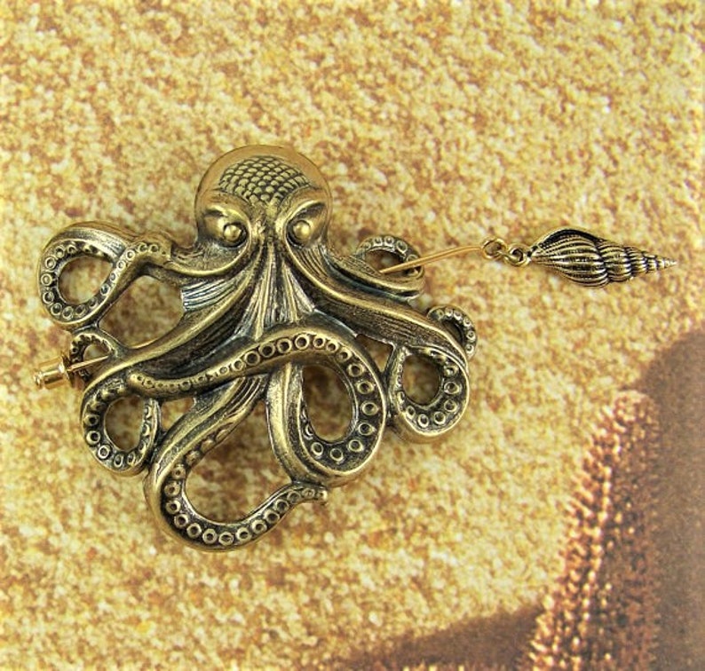 K&ouml;nnte beinhalten: Eine goldfarbene Brosche in Form eines Kraken mit einem detaillierten Design und einem Anh&auml;nger in Form einer Muschel. Die Brosche ist ein dekoratives Accessoire f&uuml;r Kleidung oder Taschen.