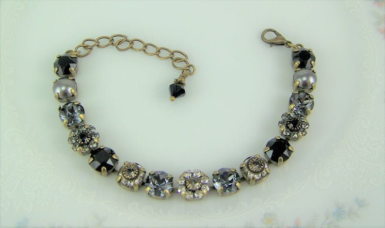 Puede incluir: Una pulsera que presenta una serie de cristales redondos y facetados en negro y gris, montados en engastes de metal color lat&oacute;n antiguo. La pieza incluye una cadena extensora color lat&oacute;n con cierre de mosquet&oacute;n y un peque&ntilde;o dije de cristal negro.