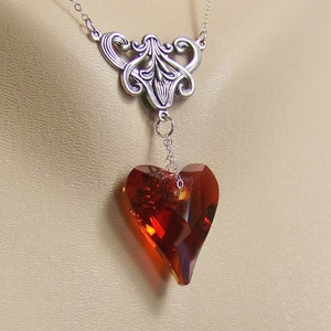 May include: A silver necklace with a delicate, ornate pendant featuring a large, faceted, red heart-shaped crystal.