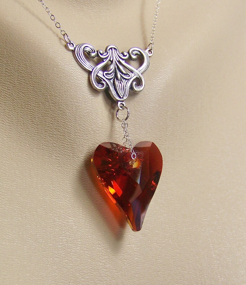 May include: A silver necklace with a delicate, ornate pendant featuring a large, faceted, red heart-shaped crystal.