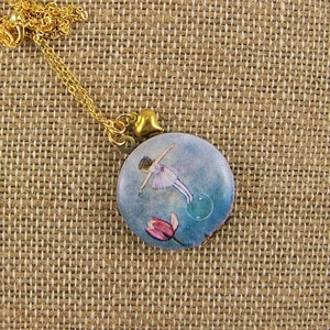 May include: A gold chain necklace with a round pendant featuring a watercolor illustration of a ballerina in a pink dress, floating above a blue and green background with a pink flower.