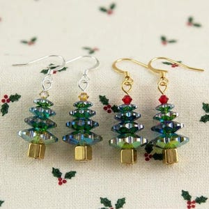 May include: Four Christmas tree earrings with silver and gold hooks. The trees are made of green and blue iridescent beads, with a gold base and a red bead at the top. The earrings are displayed on a white fabric with holly berry patterns.
