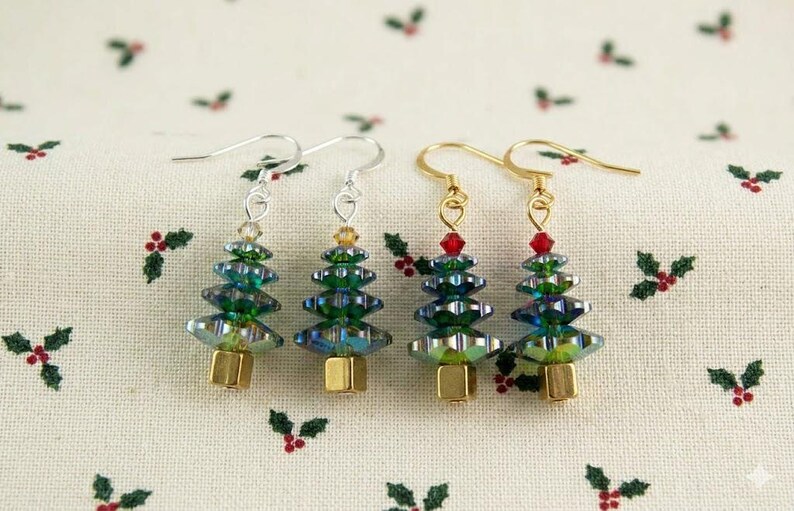 May include: Four Christmas tree earrings with silver and gold hooks. The trees are made of green and blue iridescent beads, with a gold base and a red bead at the top. The earrings are displayed on a white fabric with holly berry patterns.