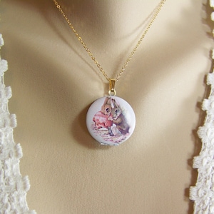May include: A gold chain necklace with a round locket featuring a vintage illustration of two rabbits. The locket has a hinged opening.