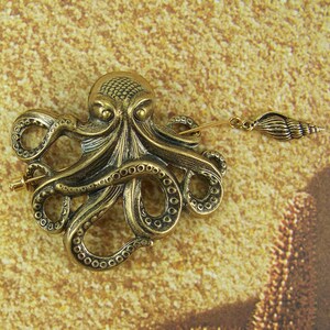 K&ouml;nnte beinhalten: Eine goldfarbene Brosche in Form eines Kraken mit einem detaillierten Design und einem kleinen Muschel-Anh&auml;nger, der an der Nadel befestigt ist.