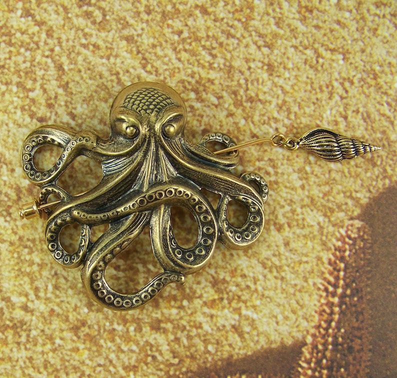 K&ouml;nnte beinhalten: Eine goldfarbene Brosche in Form eines Kraken mit einem detaillierten Design und einem kleinen Muschel-Anh&auml;nger, der an der Nadel befestigt ist.