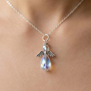 Puede incluir: Un collar de plata con un colgante de &aacute;ngel. El colgante presenta un cuerpo de cristal transparente e iridiscente en forma de l&aacute;grima, alas plateadas y una corona. El collar se lleva alrededor del cuello.