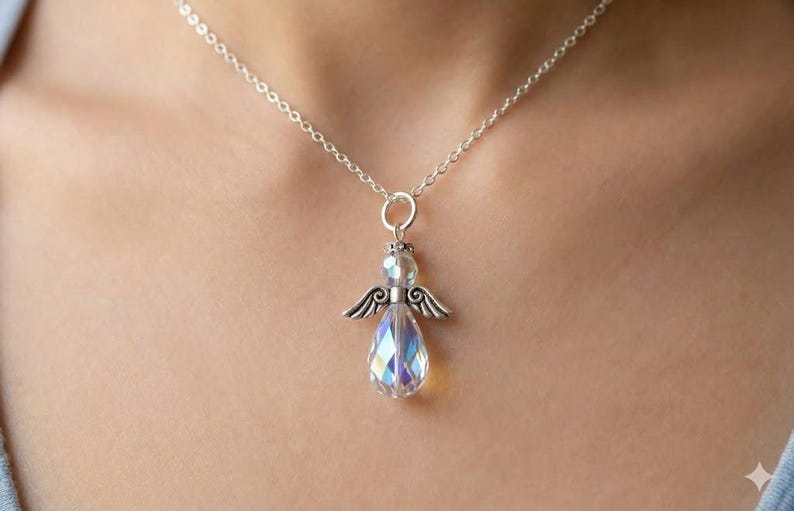 Puede incluir: Un collar de plata con un colgante de &aacute;ngel. El colgante presenta un cuerpo de cristal transparente e iridiscente en forma de l&aacute;grima, alas plateadas y una corona. El collar se lleva alrededor del cuello.